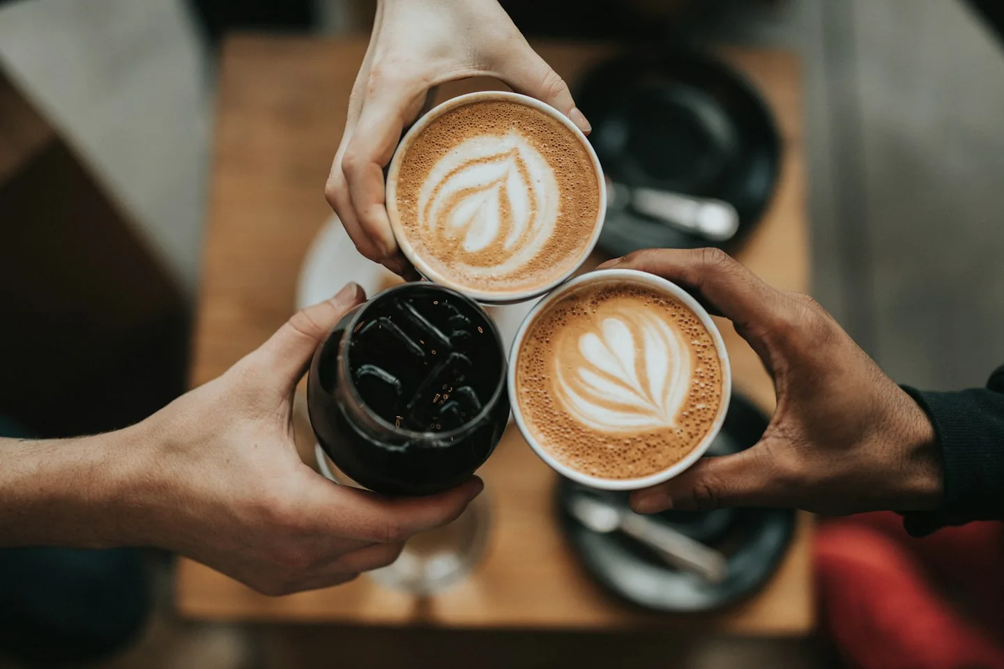 Three hands holding cups of coffee with milk art above a dark tabletop
