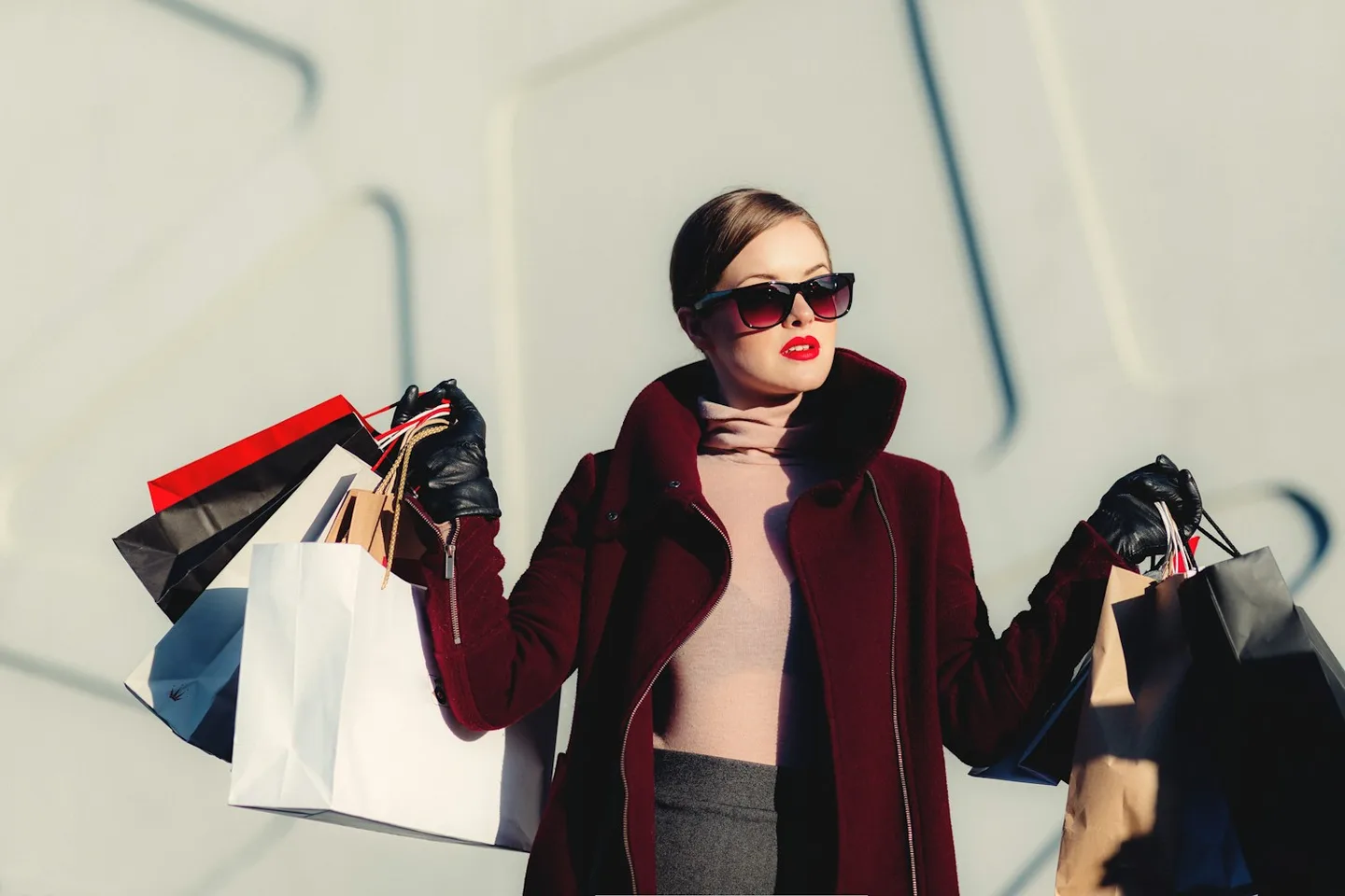 Woman in a dark red coat and sunglasses carrying shopping bags outdoors